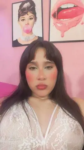 Young woman with long dark hair and pink blush wearing a white lace top in a pink room with pop art decor.
