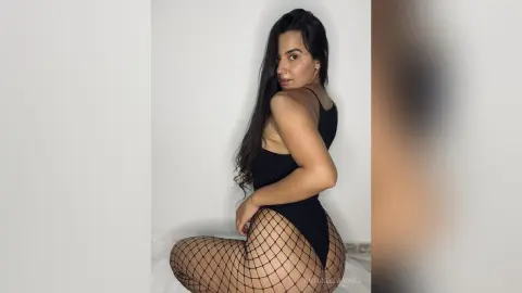 Brunette woman in a black bodysuit and fishnet tights posing against a white wall in a minimalist setting.