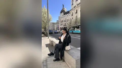 NashiroSakurai sits on a stone ledge in a black coat, white top, and grey trousers against a city street background.