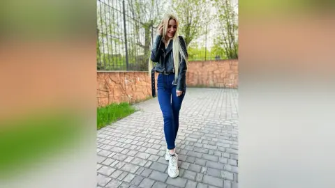Blonde woman in a black leather jacket and blue jeans standing on a stone walkway outdoors.