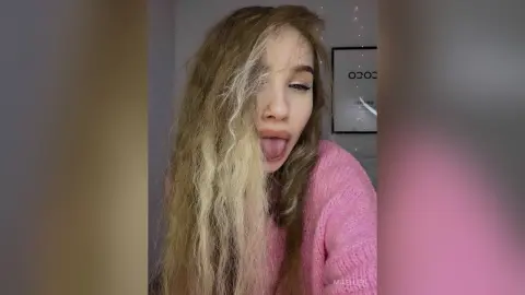 Young woman with long wavy blonde hair wearing a cozy pink sweater in a bright indoor setting.