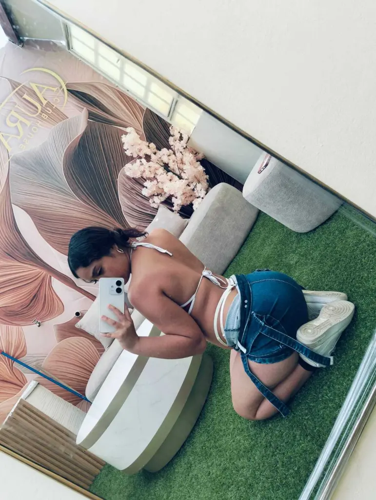 CharlotAura — Young woman in a white bikini top and denim shorts posing in a stylish salon with floral decor and green turf.