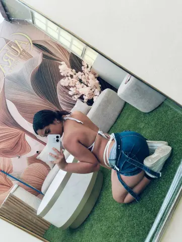 Young woman in a white bikini top and denim shorts posing in a stylish salon with floral decor and green turf.