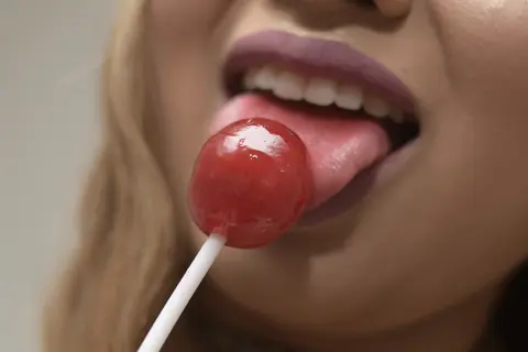Close-up of a woman with mauve lipstick enjoying a red cherry lollipop in a soft, minimalist setting.
