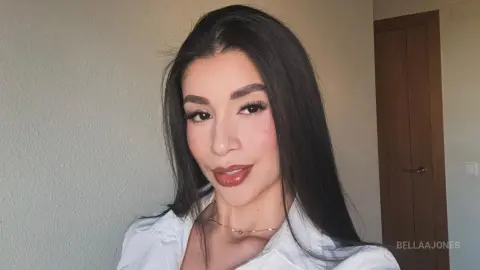 BellaaJones poses in a white shirt with her long dark hair down, looking at the camera.