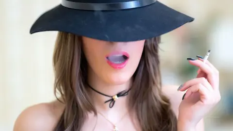 MontseFerrera wears a black wide-brimmed hat and a black choker while holding a lit cigarette near her mouth.
