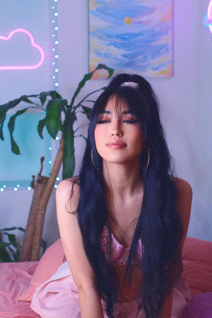 ThaliaZafirr — Young woman with long dark hair in a pink outfit sitting in a dreamy room with neon cloud lights.