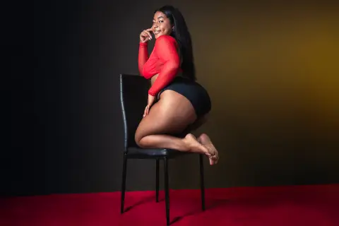 Smiling Black woman in a red long-sleeve top and black bodysuit sitting on a chair against a dark studio backdrop.