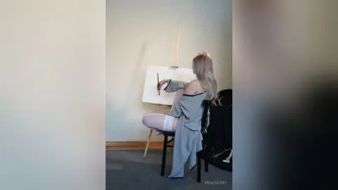 Blonde woman in a grey knit sweater painting on a canvas in a bright, minimalist studio setting.