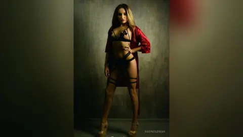Glamorous woman in black lace lingerie and red robe posing in a moody, dimly lit studio setting.