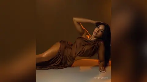 Elegant woman in a flowing brown satin dress reclining in a warm, moody studio setting.