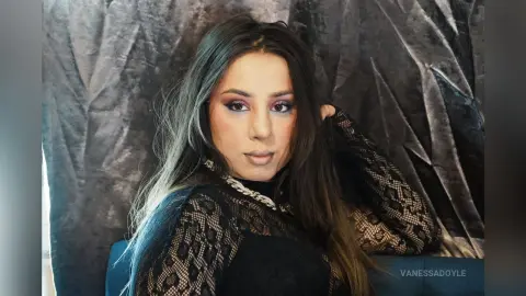 Young woman with long hair wearing a black lace top posing against a textured grey backdrop.