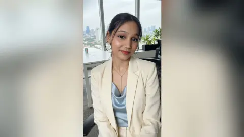 Young woman in a cream blazer and blue satin top sitting in a modern bright office setting.