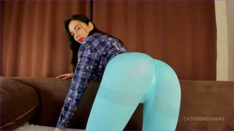 Young woman in a blue plaid shirt and light blue leggings posing on a brown sofa in a cozy room.