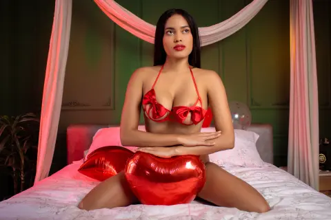 Beautiful woman in red bikini sitting on a bed with heart-shaped balloons in a romantic setting.