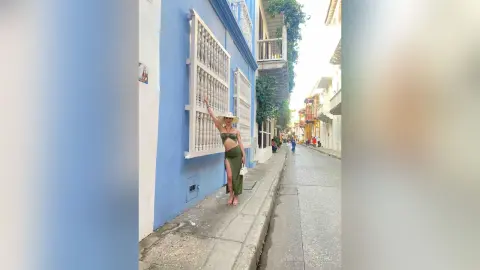 Stylish woman in an olive green bikini and maxi skirt walking along a bright blue street in a sunny coastal town.