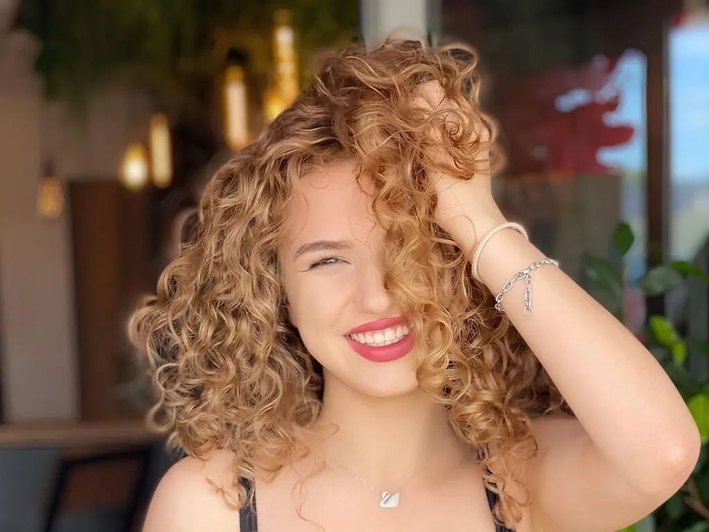 Young woman with curly hair and red lipstick wearing pearl necklace in moody pink lighting.