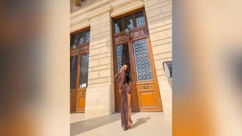 NicoleBella stands in front of large wooden doors wearing a long, form-fitting brown dress that shows her curves.