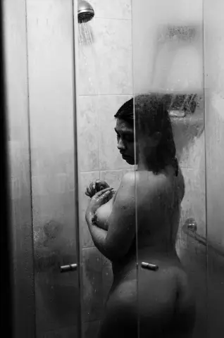 Nude woman standing in a steamy glass shower with a moody, cinematic black and white aesthetic.