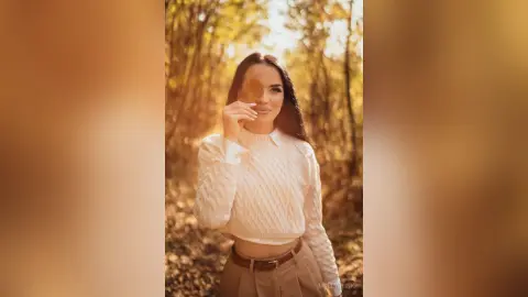 Young woman in a white cable knit sweater holding a leaf in a golden autumn forest setting.