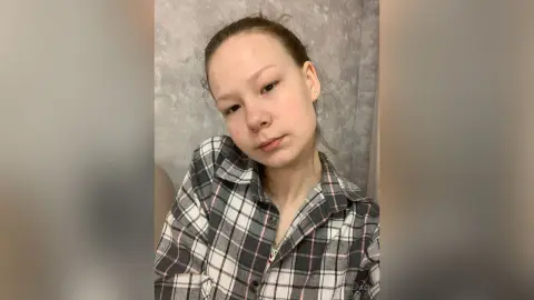 Young woman in a plaid flannel shirt sitting against a textured grey wall in a casual indoor setting.