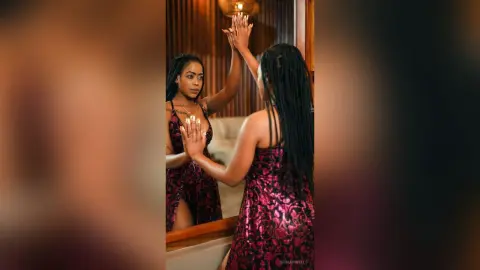 Elegant woman with long braids in a patterned burgundy dress posing confidently in a warm, moody indoor setting.
