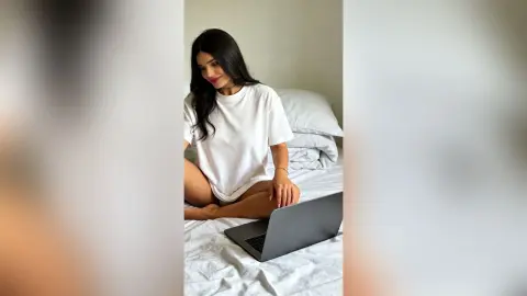 Smiling young woman in a white t-shirt working on a laptop in a bright, minimalist bedroom setting.