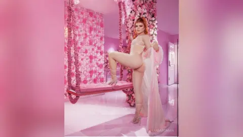 Redheaded woman in white lingerie posing on a swing in a dreamy pink floral studio setting.