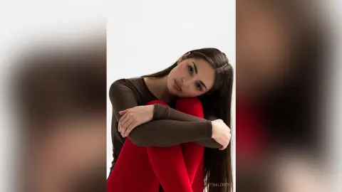 Young woman in a brown long-sleeve shirt and red leggings sitting pensively against a white background.