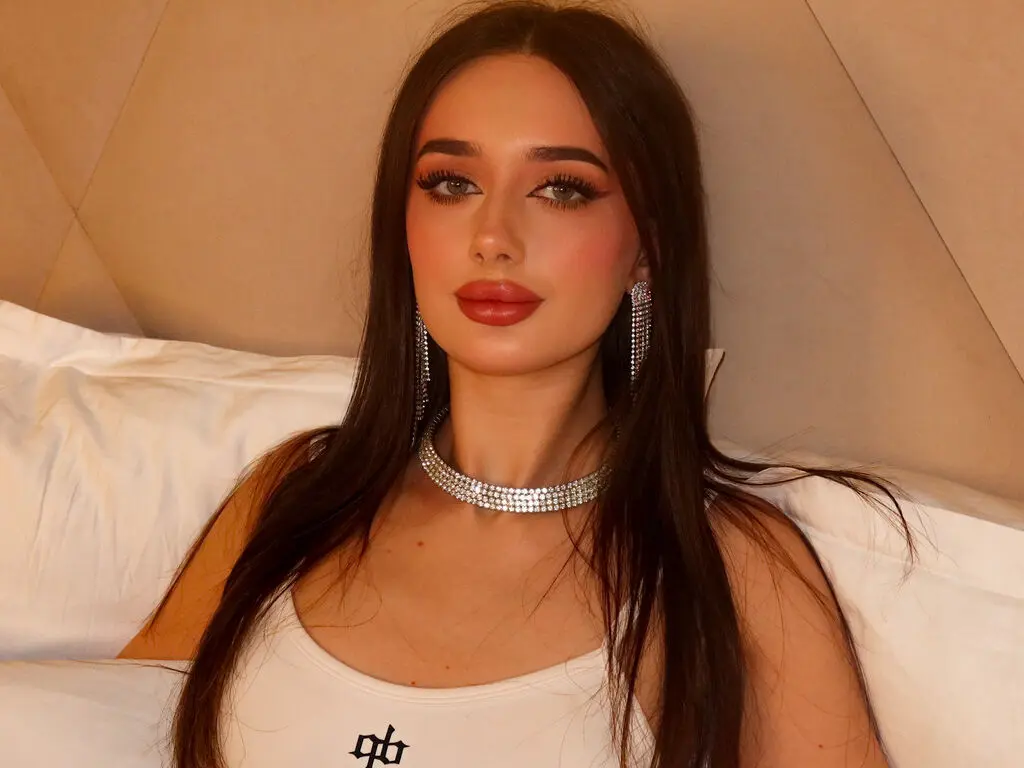 Young woman with long dark hair wearing a white tank top and silver choker in a cozy bedroom setting.