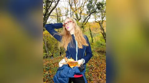 Blonde woman in a blue jacket holding autumn leaves in a sunny fall forest.