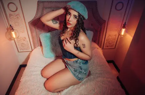 Young woman in denim shorts and blue beret sitting on a cozy bed in warm, moody lighting.