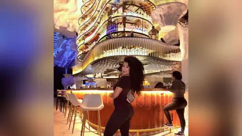 Woman in a black outfit smiling at a luxury modern bar with glowing warm lighting and tiered glass displays.