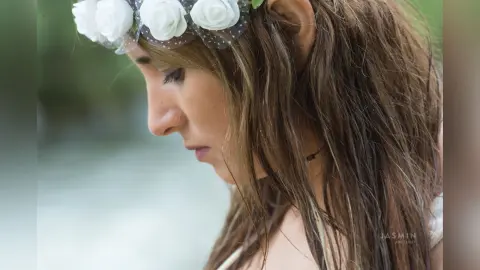 AndreaKalo looks down with her long brown hair flowing, wearing a white flower crown or headpiece.