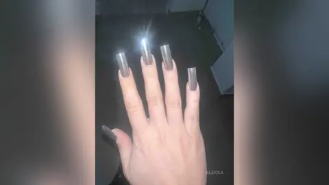 Close-up of a hand with long silver glittery stiletto nails in a dimly lit room.