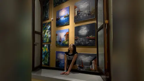 KateGale poses naked against a wall of landscape paintings, leaning forward with her hands on a ledge and looking up.