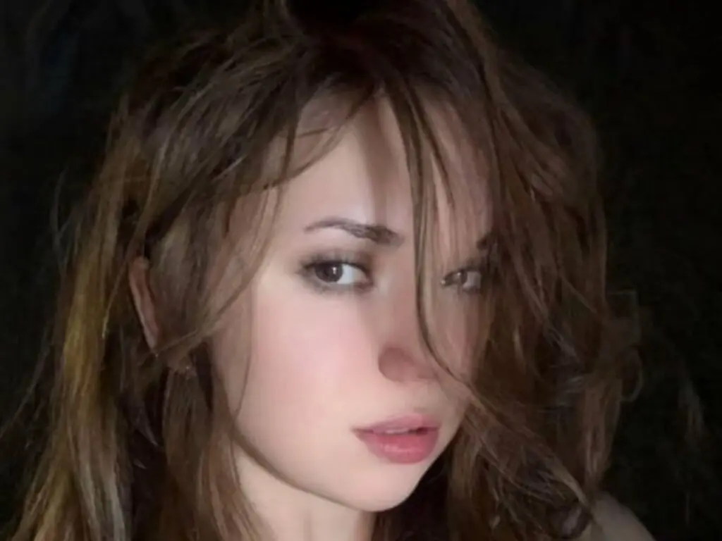Close-up portrait of a young woman with wavy brown hair and natural makeup in soft, moody lighting.
