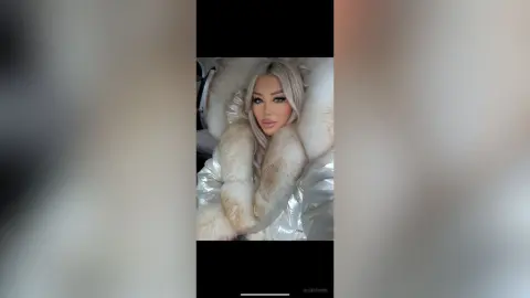 AlexaEvans poses in a shiny silver puffer jacket with a thick, light tan fur collar and long blonde hair.