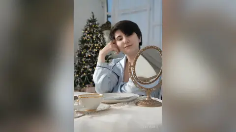 Young woman in light blue pajamas relaxing by a Christmas tree with a vintage mirror and tea cup.