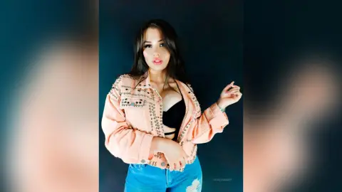 Young woman in a peach denim jacket and black crop top posing against a dark studio background.