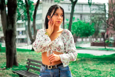 Young woman in a white floral crop top sitting on a park bench on a sunny day.