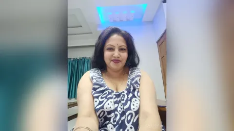 ManishaJi — ManishaJi smiles while wearing a blue and white patterned sleeveless top in her room.