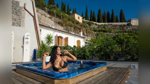MissyJolie relaxes in a blue hot tub wearing a black bikini, holding a glass to her lips against a backdrop of white buildings and green trees.