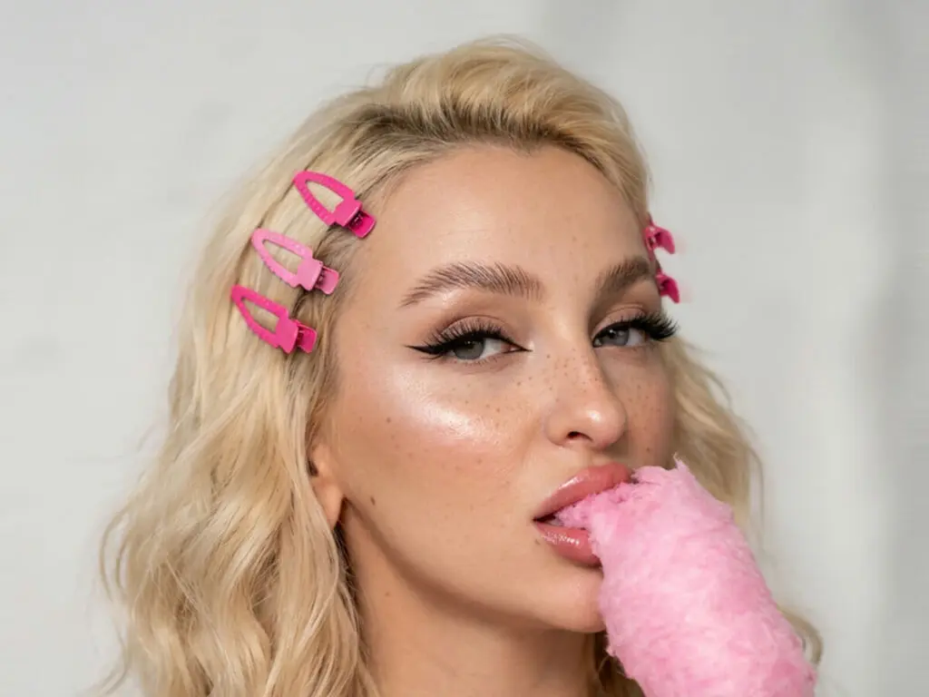 Blonde woman with pink hair clips eating pink cotton candy in a bright, playful studio setting.