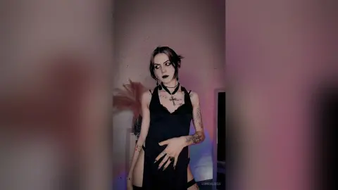 LorenLoiss poses in a black tank top and black choker, showing off her tattoos and dark makeup against a plain wall.