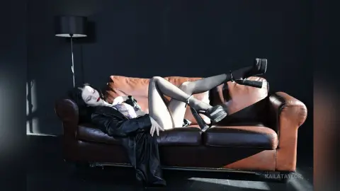 KailaTaylor reclines on a brown leather sofa wearing a black lace bra and black stockings with garters and platform heels.