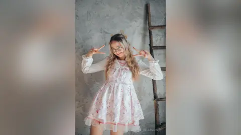 Cheerful young woman in a pink floral cottagecore dress posing with peace signs against a grey wall.
