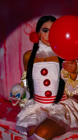 Woman in a white clown costume holding a red balloon in a spooky, moody setting.