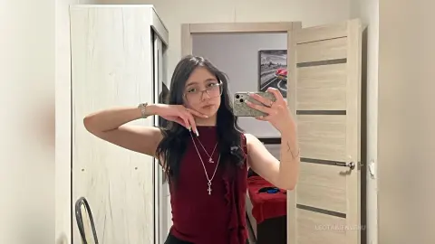 Young woman in a burgundy ruffled tank top taking a mirror selfie in a modern minimalist bedroom.