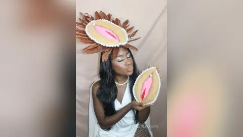 Woman in white dress holding a vulva-shaped cookie with a floral crown in a soft studio setting.